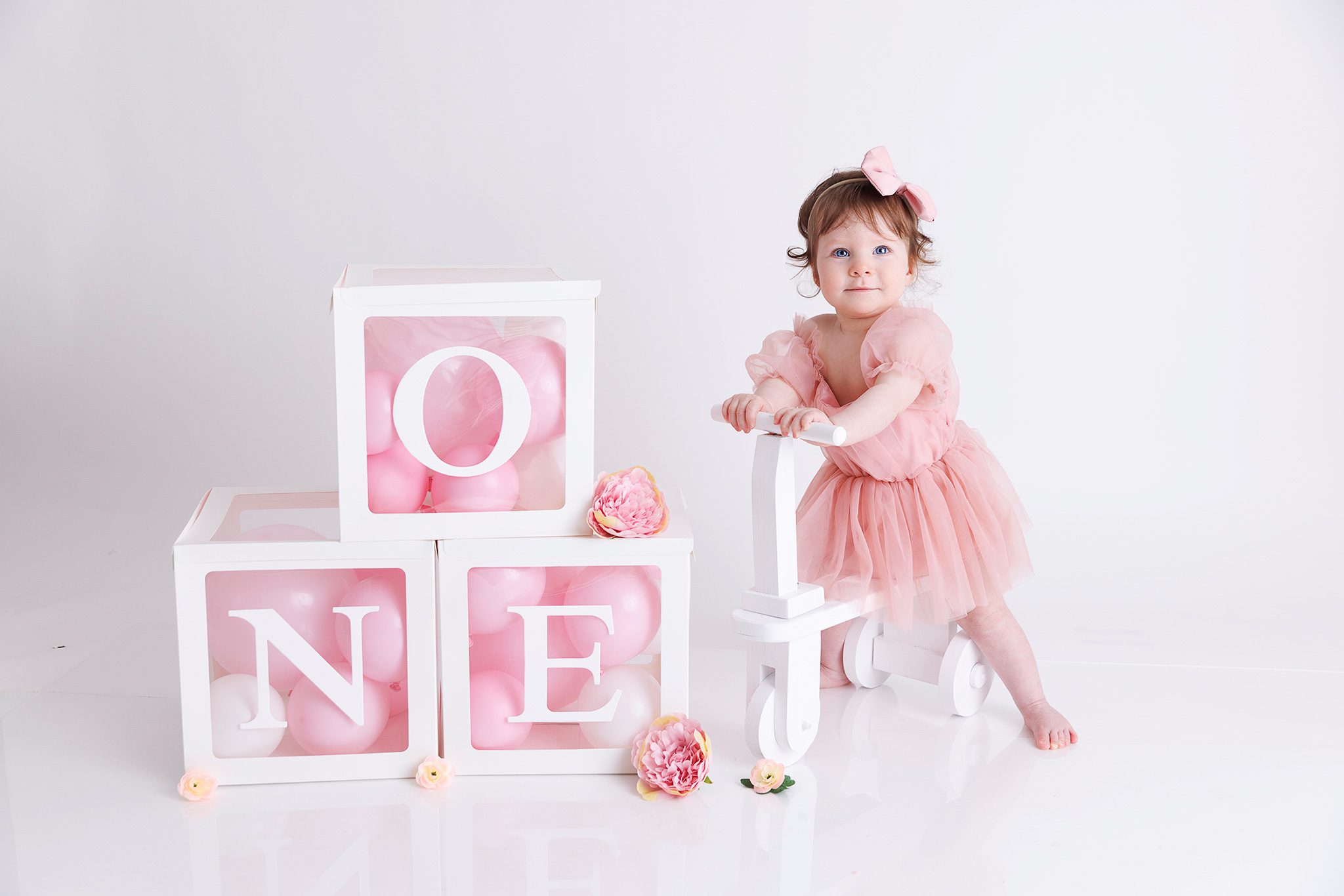 1 year old girl in pink dress posing on a little bike next to ONE boxes for a portrait photo in woking