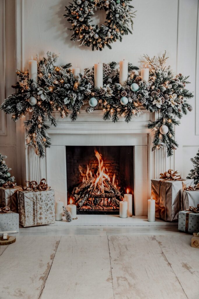 A cosy fireplace decorated with snowy garland, candles, and ornaments. A wreath hangs above, and wrapped gifts are arranged on the floor, creating a festive holiday atmosphere.